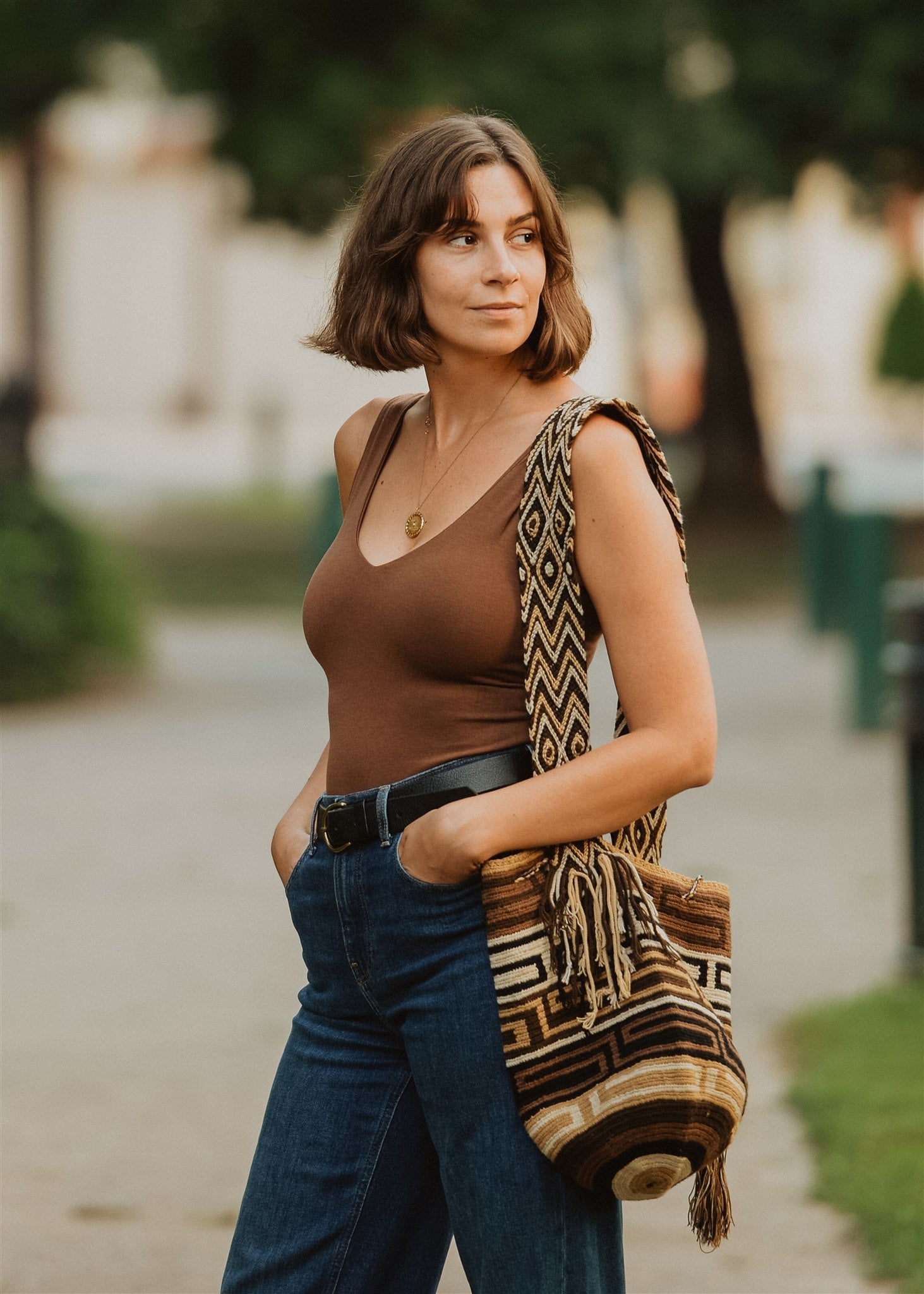 Woman standing in park hands in pockets with brown geometric pattern brown cute crossbody bag.