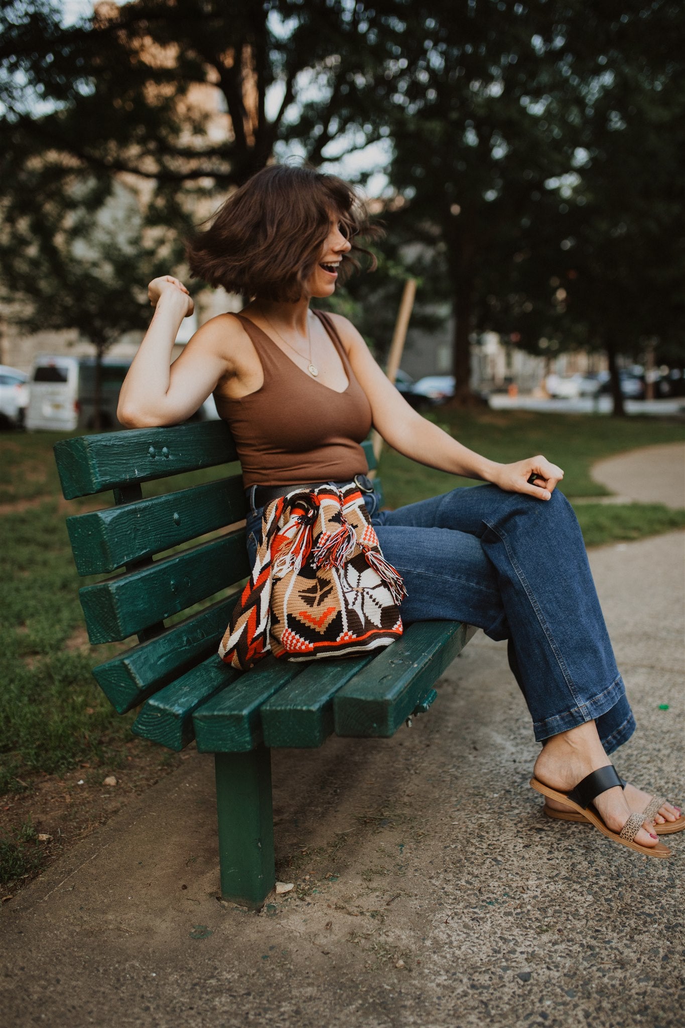 Woman smiling sitting on green park bench next to red brown white tote bag.