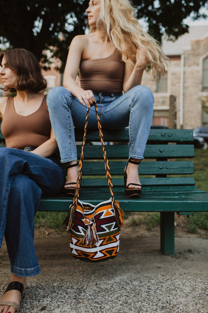 Two women wearing blue jeans brown top sitting on park bench, holding colorful bohemian tote bag.