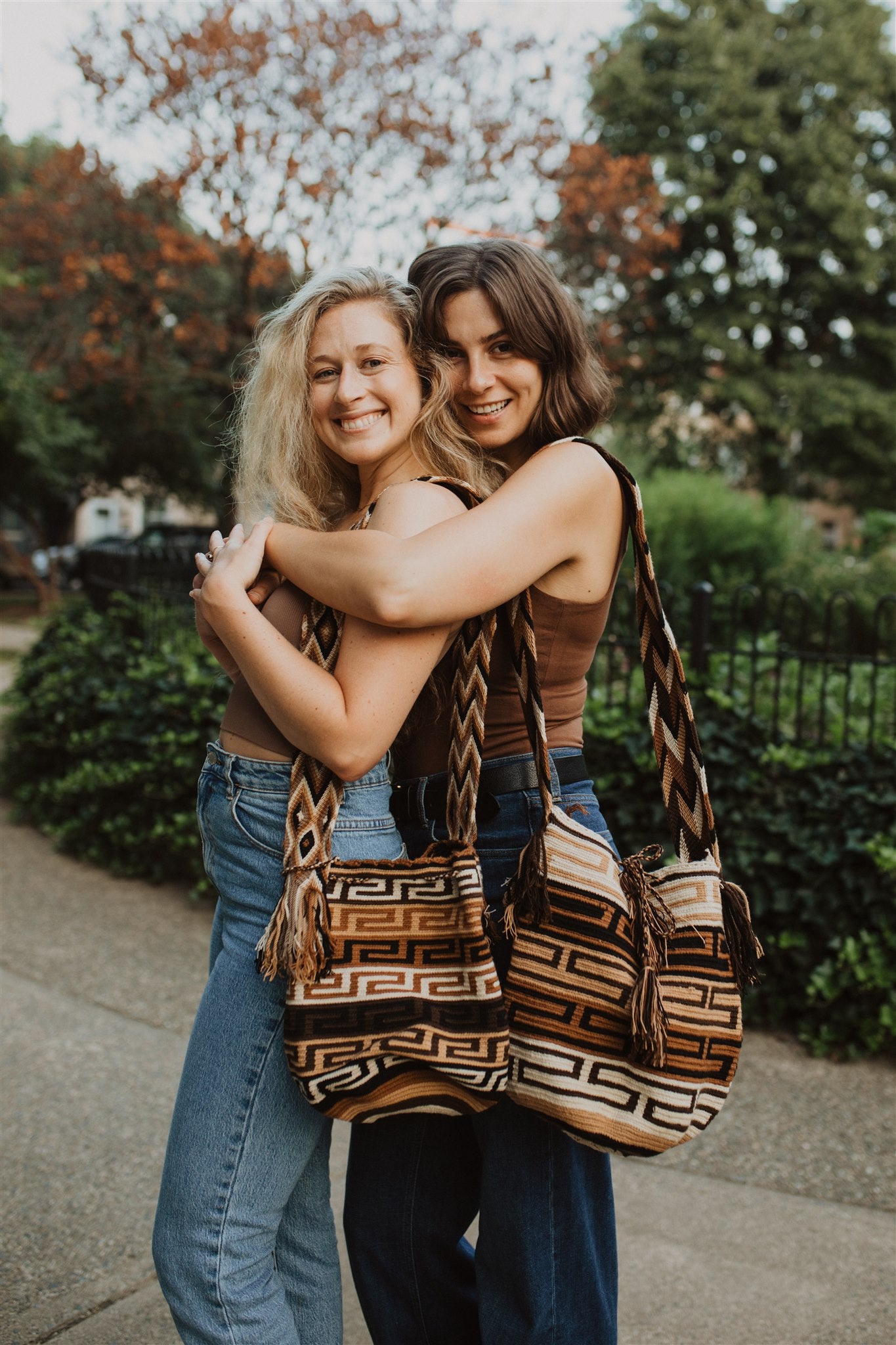 Two women wearing identical blue jean brown tops, embracing in a park wearing brown patterned boho tote bags.