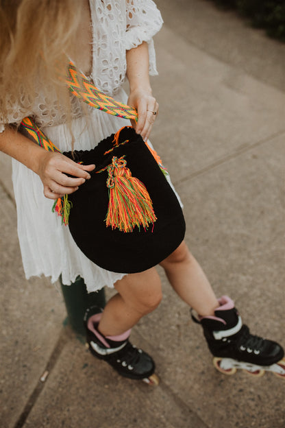 Woman in white dress and rollerblades sitting holding black boho shoulder bag with multicolored strap.