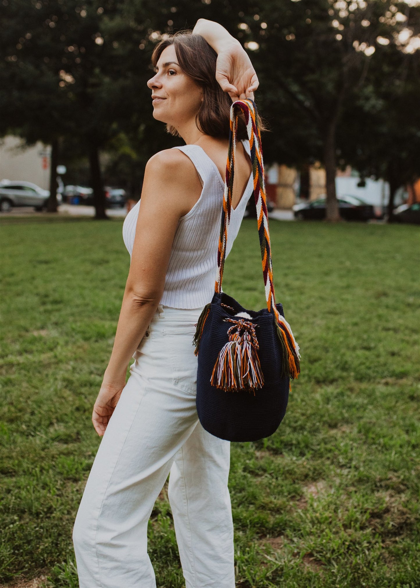 Smiling woman holding shoulder bag behind her head. Wearing white standing on grass in park.