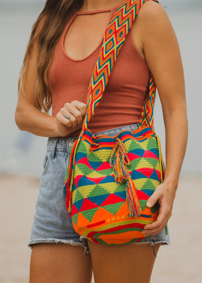 Closeup of woman holding rainbow color beach shoulder tote bag. Standing on sand water in background.