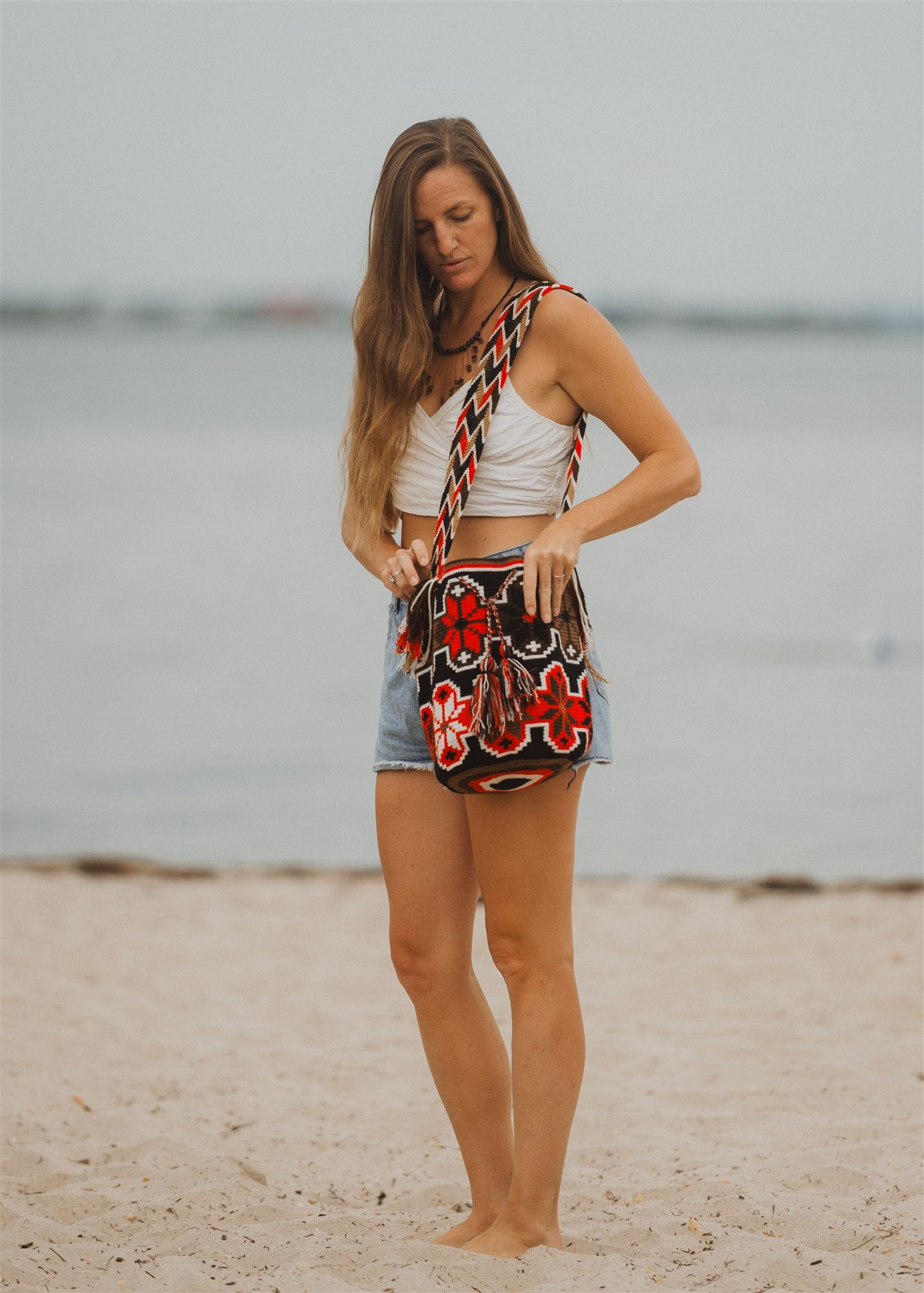 Woman standing on beach in white top and red white brown black bohemian shoulder crossbody bag.