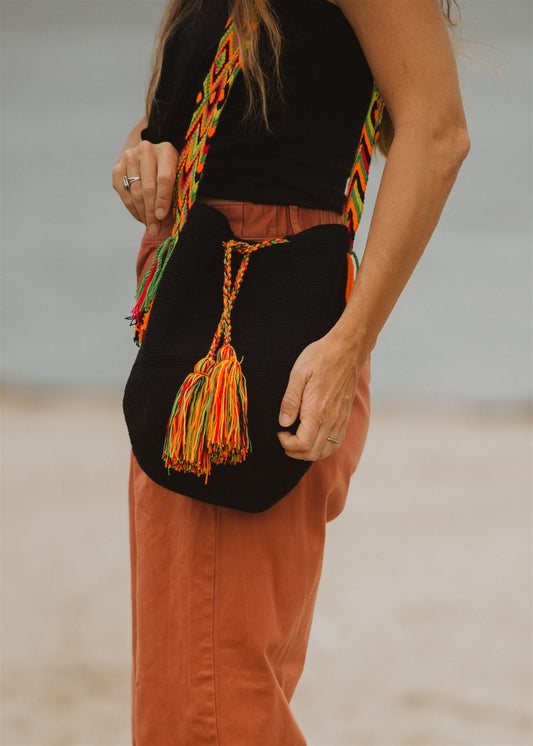 Woman on beach showing a black crochet tote bag with rainbow color strap.